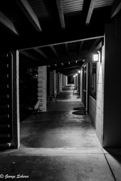 Black and white view looking down a corridor outside guest rooms at the retreat center.  The roof supports on the left and the beams overhead create a series of rectangles that diminish with distance.  Outside each room, an single lamp throws a splash of light on the concrete walkway.