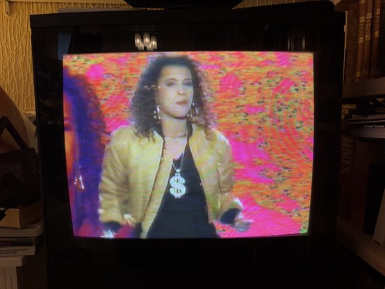 Neneh Cheery on a CRT TV in the music video for Buffalo Stance. The background is psychedelic and she is wearing a necklace with a big dollar sign. 