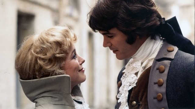A man and woman in elegant 18th‑century attire stand closely together, smiling warmly at each other. He wears a gray coat with ornate buttons, while she has curled blonde hair styled in a period fashion. The scene conveys tenderness and romance.

