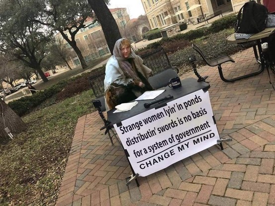 A person in medieval attire sits at a table with a sign reading, "Strange women lyin' in ponds distributin' swords is no basis for a system of government. CHANGE MY MIND." 