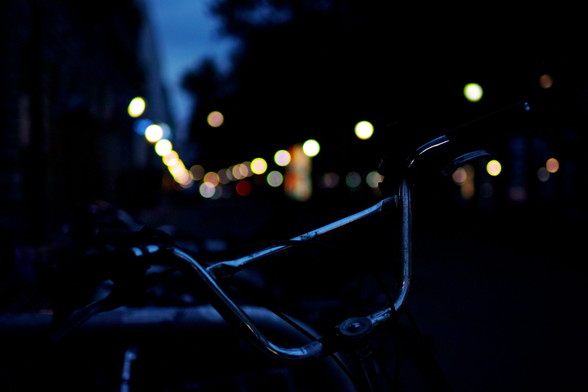 Straßenszene bei Nacht. Im Vordergrund ein Fahrrad-Lenker, im Hintergrund die Lichter der Straßenbeleuchtung als Bokeh-Bubles.