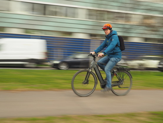 Ein Radfahrer mit Helm und Rucksack auf einem E-Bike. Der Hintergrund (parkende Autos und ein Gebäude) ist duch Mitbewegung der Kamera verwischt.