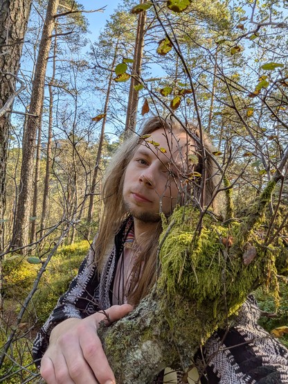 A person with long hair leans against a moss-covered tree branch in a sunlit forest, surrounded by tall trees and greenery, creating a serene atmosphere.