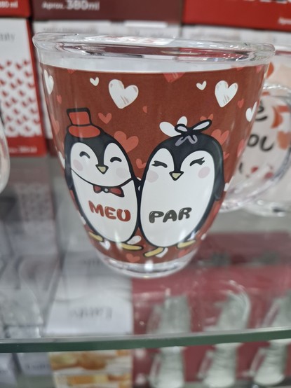A imagem mostra uma caneca de vidro transparente com uma ilustração de dois pinguins. O fundo da caneca é de cor marrom com vários corações brancos e vermelhos espalhados. Os pinguins são desenhados de forma cartoon, com um pinguim usando um chapéu vermelho e o outro com um laço preto. O pinguim com o chapéu tem a palavra "MEU" escrito em seu peito, enquanto o outro pinguim tem a palavra "PAR" escrito. A caneca está em uma prateleira de vidro, com outras canecas visíveis ao fundo, sugerindo que…