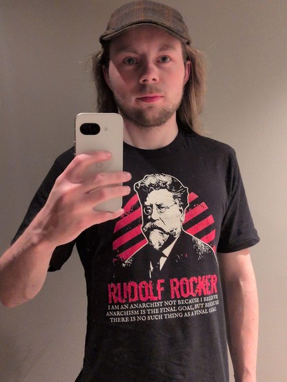 A young man with long, light blond hair and a brown tweed cap takes a selfie, holding a white phone in his left hand. He wears a black t-shirt with a portrait of Rudolf Rocker with his quote "I am an Anarchist not because I believe Anarchism is the final goal, but because there is no such thing as a final goal." displayed underneath. The background is a plain gray wall.
