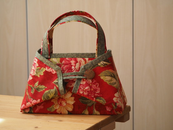 Eine Handtasche aus Stoff, der Stoff hat auf rotem Hintergrund Blütenmotive. Das Design der Tasche ist eine Art Faltung.