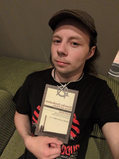 A person wearing a brown hat and black shirt holds a book titled "Anarcho-Syndicalism: Theory and Practice." They have a neutral expression, sitting on a green sofa.