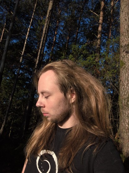 A person with long hair is standing in a sunlit forest, eyes closed, wearing a black shirt with a circular Debian design. Tall trees and deep greenery surround them.