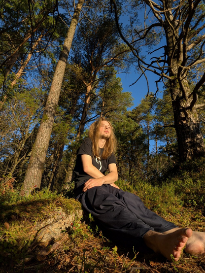 A person with long hair, wearing a black shirt with a spiral Debian design, sits barefoot in a sunlit forest. Surrounding trees and blue sky convey tranquility.