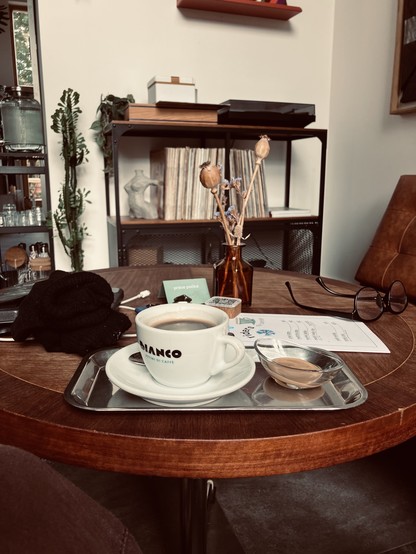 A cozy coffee shop scene featuring a wooden table with a cup of coffee on a tray, accompanied by a small dish. Nearby, there are dried flowers in a vase, a pair of glasses, a menu, and a dark sweater. In the background