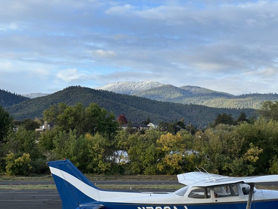 A snow-whitened mountain among an panorama of evergreen covered mountains, behind tree-covered buidings. The foreground is a small airport runway showing the top of a small plane 