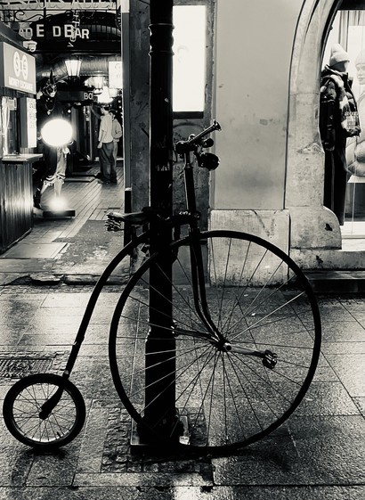 An old-fashioned penny-farthing bicycle is leaning against a street pole on a dimly lit street. The scene is captured in black and white, showcasing reflections on the wet pavement and a pedestrian in the background.