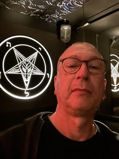 A person with glasses is posing for a selfie in a dimly lit room featuring a large illuminated pentagram symbol on the wall. The setting appears to have a dark and moody aesthetic.
