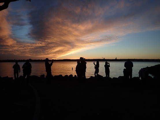 View of a sunset, looking across San Diego Bay, with Coronado Island in the background, with orange clouds and blue sky.  The foreground is dark, with silhouettes of people taking photos of the sunset, each other, and themselves with their phones.