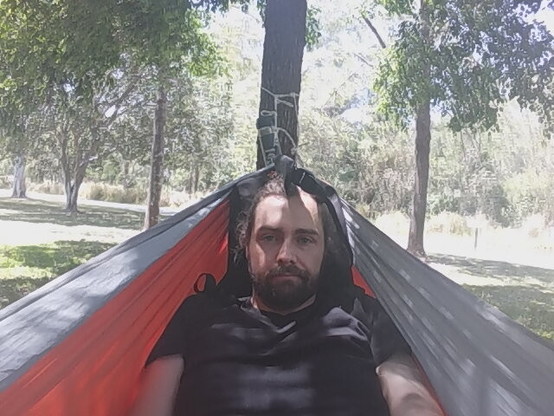 Man in an orange and gray hammock, head resting on a laptop bag and shoulders looking a bit squeezed together, but comfortable nonetheless. 