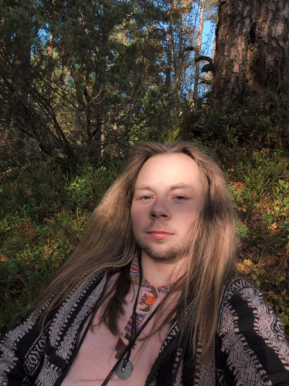A person with long hair sits in a sun-dappled forest, wearing a patterned sweater and necklace. The scene conveys tranquility and connection with nature.