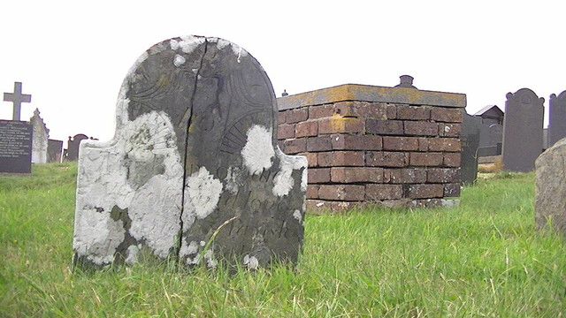 a pretty slate head stone that has split down the middle. idk it looks almost like art nouveau with lashings of white lichen. cursive writing. cant make out all of what it says tho. 