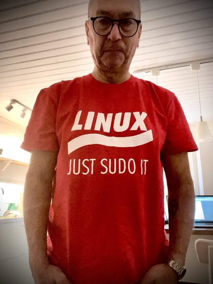 An individual is wearing a red t-shirt with the text "LINUX JUST SUDO IT." The background shows a kitchen setting with various items. The person appears to have glasses and is standing with a neutral expression.