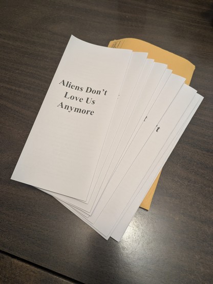 Black and white pamphlets resting over a brown envelope on a table. The front of them read "Aliens Don't Love Us Anymore".