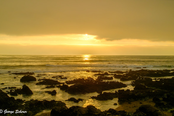 As it descends at the end of the day, the sun is emerging from low clouds, illuminating the waves on the ocean. There is a rocky beach in the foreground. The sun's illumination gives everything in the scene a golden glow.