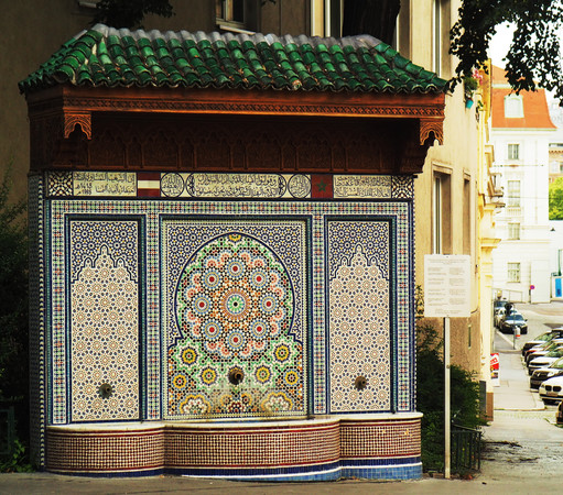 Ein Brunnen mit 3 Wasserauslässen, die Rückwand ist mit einem bunten Mosaik gekachelt, über dem Mosaik eine Inschrift in arabischer Schrift.