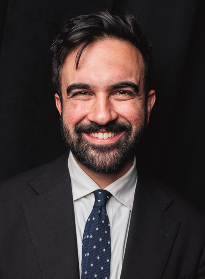A man with short dark hair and a full beard smiles warmly at the camera. He’s wearing a dark suit, white dress shirt, and navy blue tie with small white dots. The background is black.