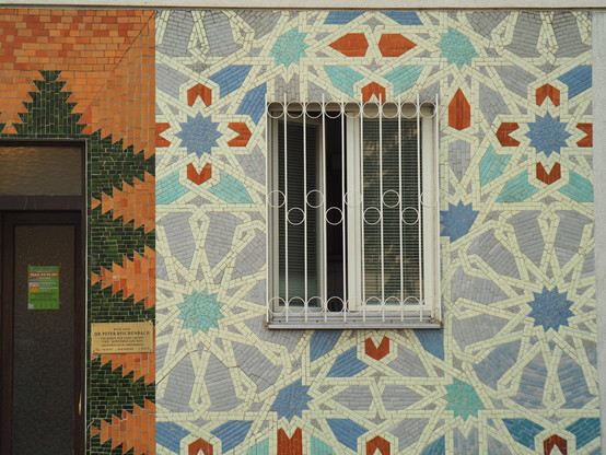 Auf einer Fassade ein islamisches Sternenmuster und dazwischen ein Fenster mit Gitter davor. Am linken Bildrand noch ein Teil einer Tür.