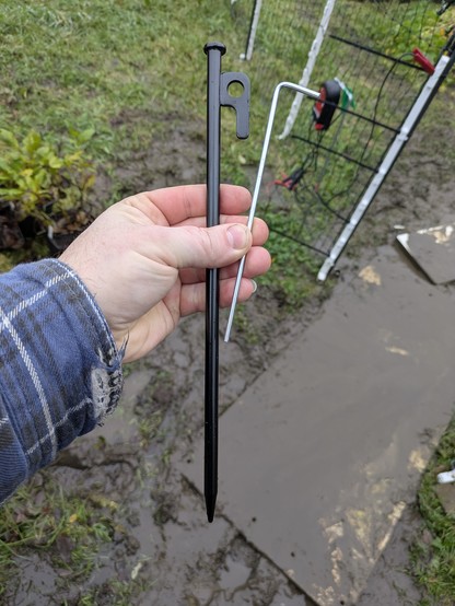 A pathetic little galvanized steel wire tent peg next to a massive 12"/30cm x 1/2"/10mm powder-coated steel sabot with a welded plate hook