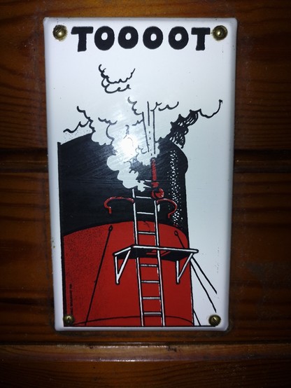 se ve una chapa clavada a una pared de madera. en la chapa hay una chimenea de un barco que está haciendo sonar el silvato y está escrito "toooot"