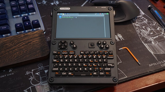 The Clockwork uConsole kit fully assembled sitting on a desk beside a keyboard and a mouse on a NASA-themed desk mat.  The device is booted into Linux with a console terminal window open.