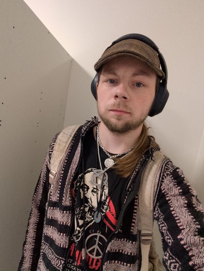 A person with a light beard and long hair pulled back is taking a selfie indoors. They are wearing over ear headphones, a brown cap, layered necklaces, a patterned hoodie, and a backpack. Their T shirt features a graphic portrait and a necklace with the peace symbol. The background is a plain light wall.