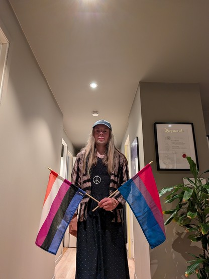 A gender-fluid person stands in a softly lit hallway holding two pride flags, one gender-fluid and one bisexual. They are wearing a green cap and a black dress. Framed art and a potted plant are visible in the background, adding a cozy and calm atmosphere.