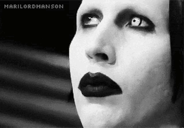 A black-and-white close-up of a person wearing dramatic gothic makeup, including dark lipstick, heavy eyeliner, and light contact lenses. They look upward with an intense, expressionless gaze.