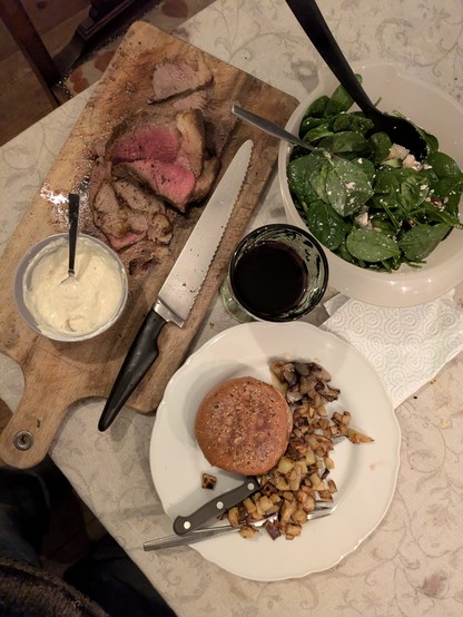 A cut of roasted beef, sauce made with fresh horseradish from the garden, a seedy bun, fried potato cubes, spinach salad, and red wine