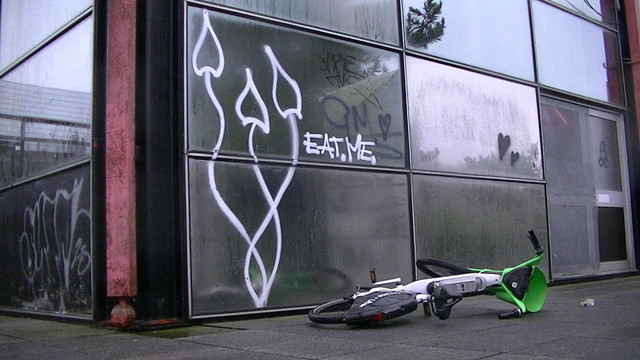 liberty caps graffiti on glass paneled derelict  building with caption "EAT.ME." an uber bicycle or whatever lays on the discarded pavement