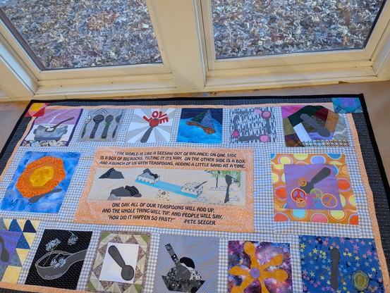 A quilt of different styles based on the theme of "spoons". In the center is a quote from Pete Seeger. "The world is like a see-saw out of balance. On one side is a box of big rocks, tilting it its way, and on the other side is a box, and a bunch of us with teaspoons, adding a little sand at a time. One day all our teaspoons will add up, and the whole thing will tip, and people will say 'How did it happen so fast?'"