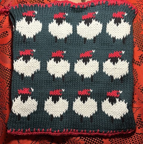 A knitted pot holder featuring a pattern of white sheep with black faces, each wearing a red pixie hat. The background is dark green, and the edges are finished with a red trim. The square is placed on a textured red lace surface.