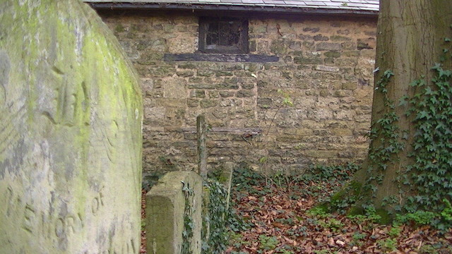 looking along a row of grave stones to a small filthy window, cobwebs and all a top of the wall of a stone outbuilding. you can see there used to be an even bigger window underneath the small window, lintel still remains but rest has been filled in with stone.