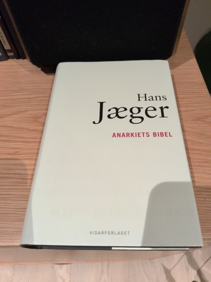 Cover of the book "Anarkiets Bibel" by Hans Jæger, featuring bold black and red text on a white background, placed on a wooden shelf.