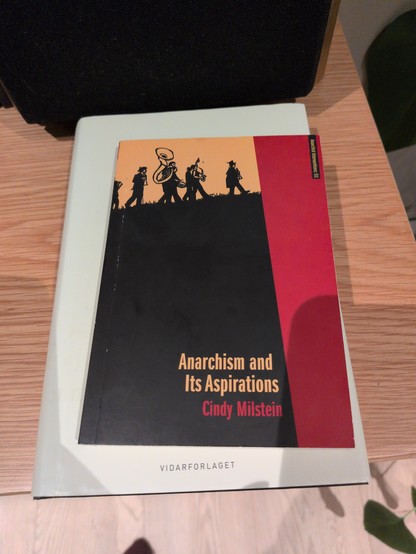 Cover of "Anarchism and Its Aspirations" by Cindy Milstein on a table. The cover features silhouettes of people walking and playing instruments.