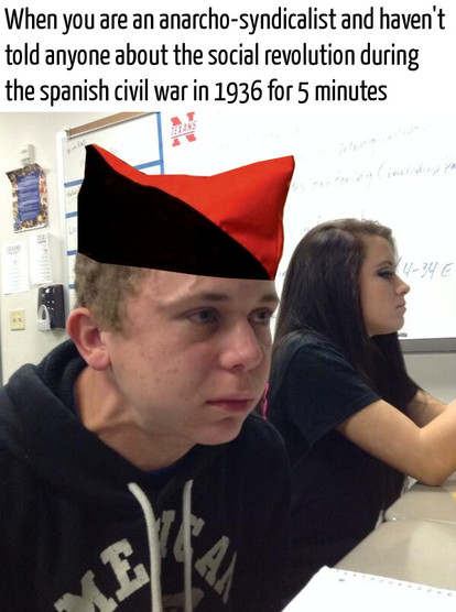 A meme-style photo of a teenage boy sitting in a classroom, staring intensely while wearing a digitally added red-and-black anarchist cap, another student sits beside him. White text at the top reads, When you are an anarcho-syndicalist and haven’t told anyone about the social revolution during the Spanish civil war in 1936 for 5 minutes.