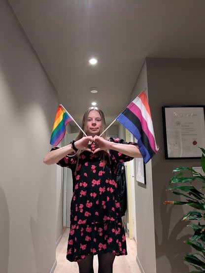 A person standing in a hallway, wearing a black dress with pink floral patterns and dark tights. They hold two small flags, one rainbow pride flag and one pink, purple, blue, black, and white gender-fluid flag, while forming a heart shape with their hands in front of their chest. Overhead lights illuminate the space, with framed art and a plant visible along the hallway walls.