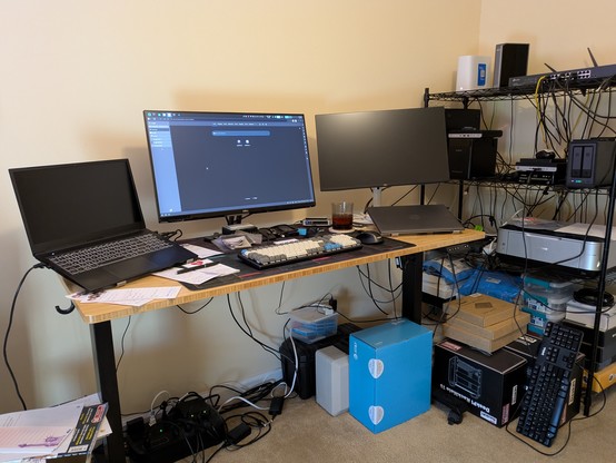 A busy home lab and developer workspace featuring a wooden desk with a laptop on a stand, two monitors, and a mechanical keyboard. To the right, a black wire shelving unit is stacked with networking hardware, including a network switch and a compact two-bay NAS. The setup is filled with visible cabling, a printer, and several tech equipment boxes on the floor, including a DeskPi RackMate box, reflecting a functional and active technical environment.
