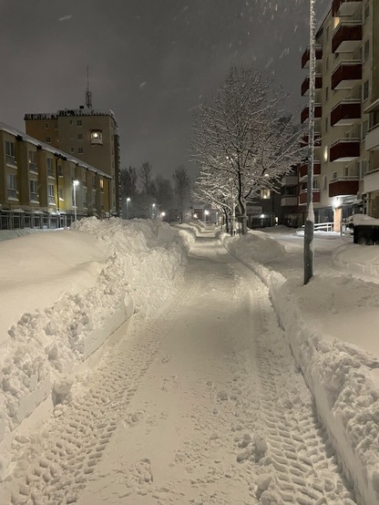 A snowy urban scene at night, featuring illuminated apartment buildings, a snow-covered pathway with tire tracks, and trees dusted with snow. Flakes are visibly falling, creating a serene winter atmosphere.