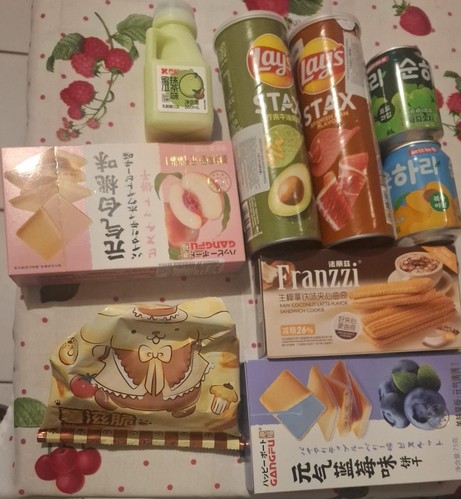 Uma variedade de itens de lanche e uma bebida estão dispostas sobre uma superfície de tecido com um padrão vermelho-laranja e verde folha, adornados por listras vermelhas. No lado esquerdo, há uma pequena garrafa de bebida verde claro “蜜桃味” (sabores de manga) (500ml), uma caixa rosa com o rótulo “五仁桃白芝麻” (manga, sementes de morango, cacau) com imagens de manga, e uma caixa amarela com um personagem cartoon e texto “喜滋脆” (feliz e crocante). No centro e à direita, há duas garrafas Lays Stax (“STA…