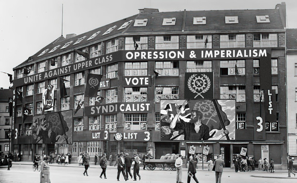 Black-and-white historical photograph of a large urban building completely covered in political banners and murals promoting syndicalism, including slogans like Unite against upper class oppression and imperialism, vote syndicalist, and list 3, with workers depicted raising flags and crowds of pedestrians walking past on the street below, suggesting an election campaign staged in a strongly working-class, revolutionary aesthetic.