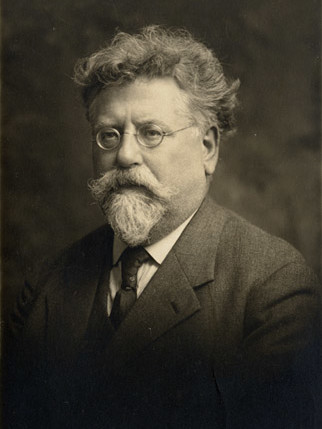 Sepia-toned portrait of an older man with wavy, graying hair, round glasses, and a thick mustache and pointed beard, wearing a dark suit, white shirt, and tie, seated and turned slightly to the side against a soft, studio-style background.