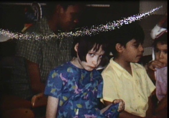 Grainy, low-light image of several young children seated closely together indoors; one child with pale skin and dark, chin-length hair looks downward with a guarded expression while wearing a patterned blue dress, sitting beside another child in a light-colored shirt, with other figures partially visible in the background.