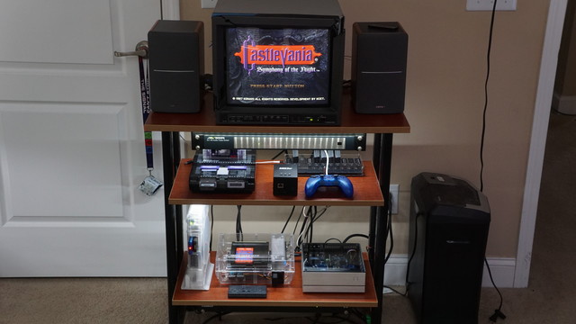 A JVC PVM sitting between two large-ish speakers on top of a printer cart.  The shelf below has a SNES sitting beside a smol MiSTer FPGA.  Beside that is a charging PS1 controller and a SCART switch.  The bottom shelf holds a PS1 console in a clear acrylic case, an NES in a clear acrylic case, and another MiSTer that's actually hooked up.