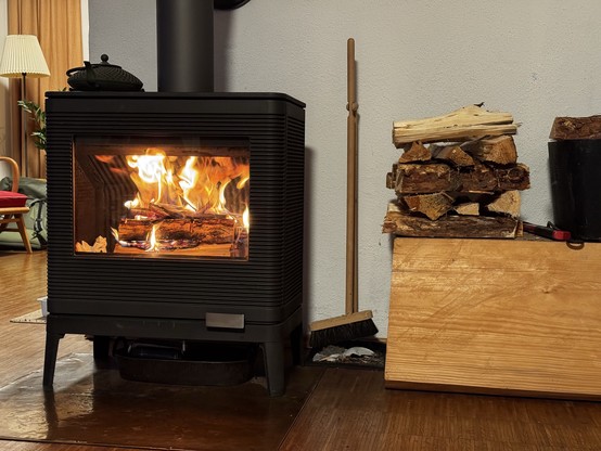 Auf dem Bild ist eine gemütliche Wohnraumszene zu sehen. Im Mittelpunkt steht ein schwarzer Holzofen mit Sichtfenster, in dem ein lebhaftes Feuer brennt. Die Flammen umschließen mehrere Holzscheite und tauchen den Raum in ein warmes, orangefarbenes Licht. Auf dem Ofen steht eine dunkle, gusseiserne Kanne.

Rechts neben dem Ofen befindet sich ein Holzlager: Auf einer hölzernen Kiste sind mehrere gespaltene Holzscheite ordentlich aufgestapelt. Davor lehnt ein Kaminbesen an der Wand, daneben ist e…